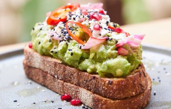 Thick-cut sourdough toast piled high with smashed avocado, feta, sliced chili, pomegranate seeds, and sesame seeds.