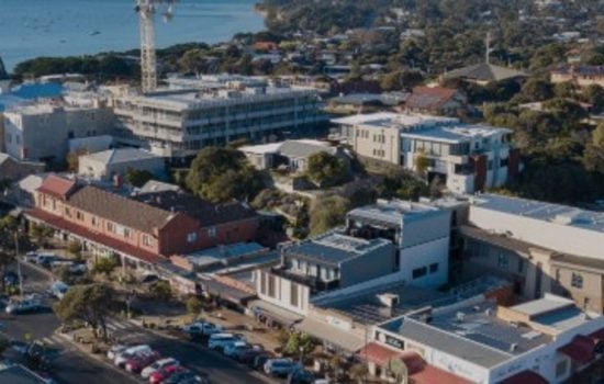 Aerial view of a coastal town featuring buildings, parked cars, and a blue ocean bay in the background.