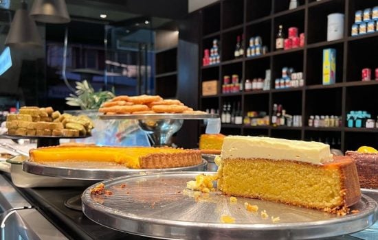 Bakery counter displaying sliced cakes, tarts, and pastries on metal stands in front of dark shelving.